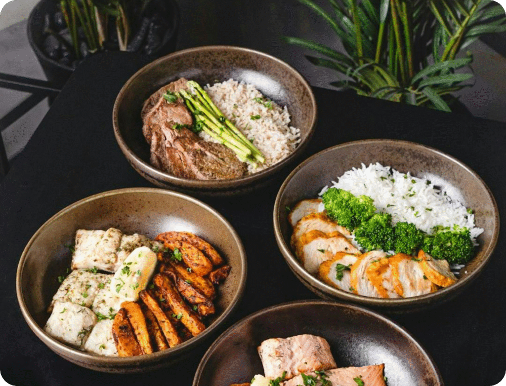 Close-up of four healthy meal prep bowls featuring grilled meats, rice, vegetables, and herbs on a black table.