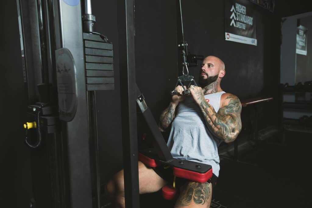 Tattooed man performing seated cable row in gym with Higher Standards Lifestyle banner in the background.