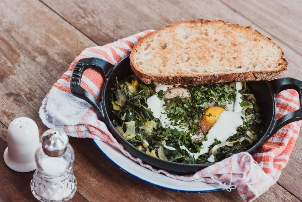 Breakfast skillet with eggs, sautéed greens, herbs, and a slice of toasted bread served in a cast iron pan.